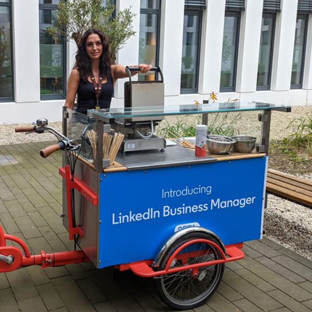 ine Frau steht hinter einem mobilen Wafflebike, das für das LinkedIn Business Manager Programm wirbt. Das Wafflebike ist in kräftigem Rot und Blau gestaltet und mit einem Waffeleisen ausgestattet. Die Frau, mit dunklen Haaren und einem schwarzen Top, bedient das Waffeleisen, während sie freundlich in die Kamera blickt. Im Hintergrund ist ein modernes Gebäude mit weißen Säulen.