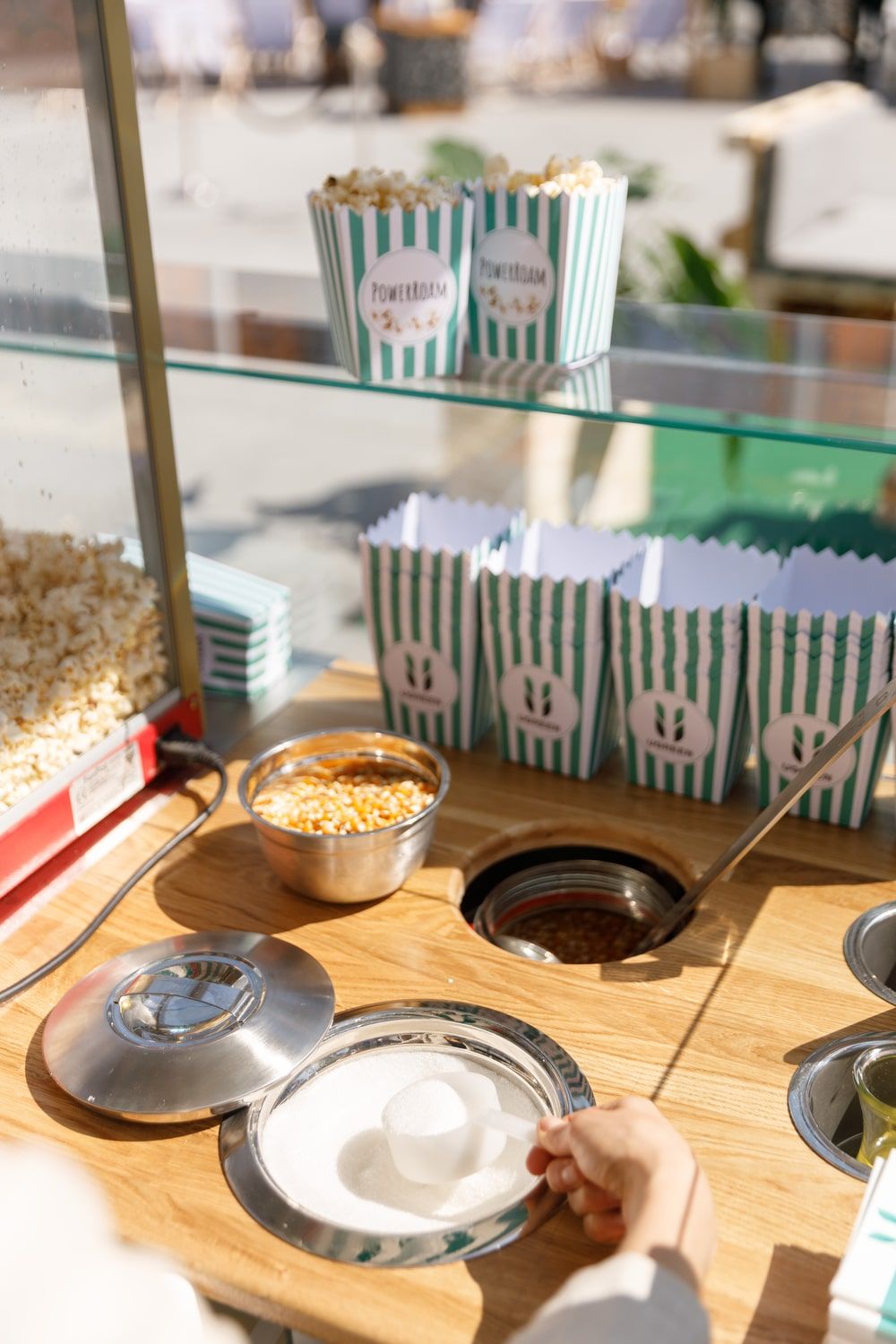 Popcornstand mit gebrandeten, grün-weiß gestreiften Popcorntüten und einer Hand, die Zucker entnimmt.