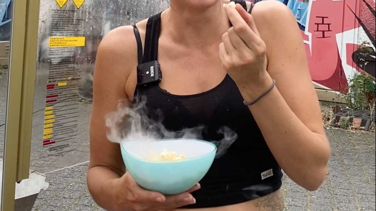 Eine Frau mit einem Lächeln im Gesicht hält eine Schüssel mit dampfendem Popcorn. Sie trägt ein schwarzes Top und steht vor einem farbenfrohen, urbanen Hintergrund mit Graffiti.