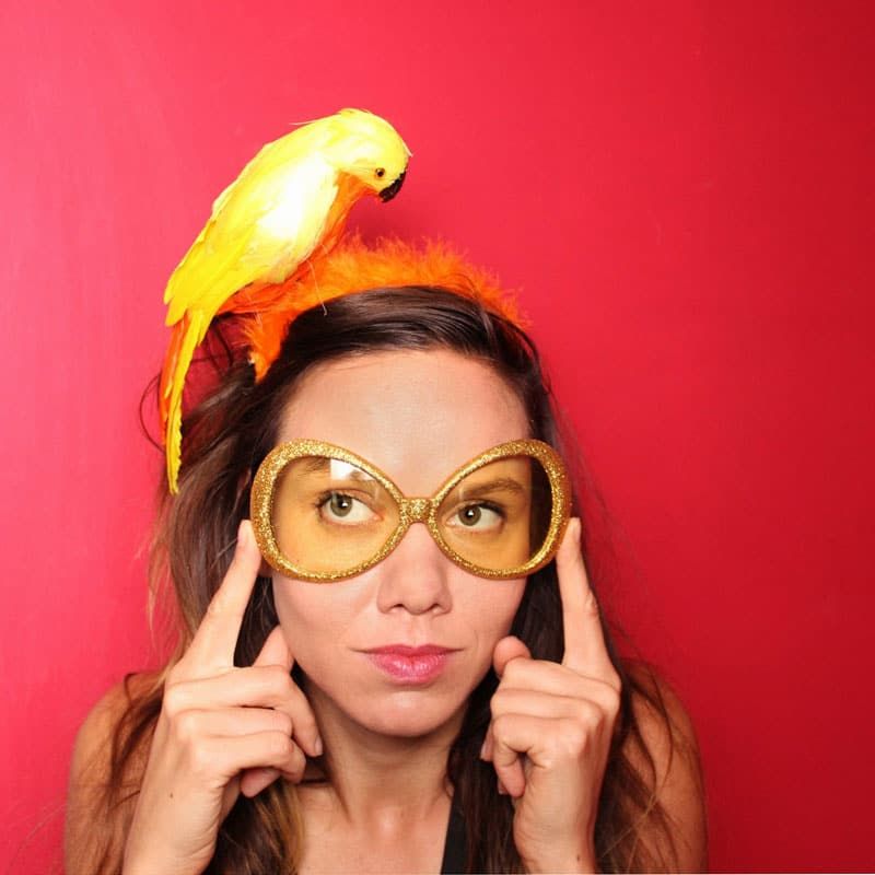 Grit trägt witzige Fotobox-Accessoires wie eine gelbe Glitzerbrille und ein Haarband mit einer Vogelattrappe vor einem roten Hintergrund bei einem Event in Düsseldorf.