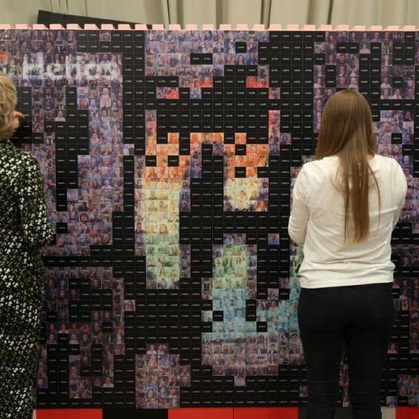 Das Bild zeigt eine große photo mosaic wall, die aus vielen kleinen Fotos besteht. Zwei Personen stehen vor der Wand und bringen Fotoaufkleber an, um das Mosaik zu vervollständigen