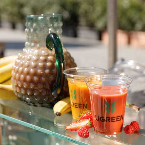 Zwei Smoothies in gebrandeten Bechern mit dem Logo von UGREEN stehen neben einer dekorativen Glaskaraffe auf einem Tisch. Im Hintergrund sind Bananen und weitere Früchte zu sehen.