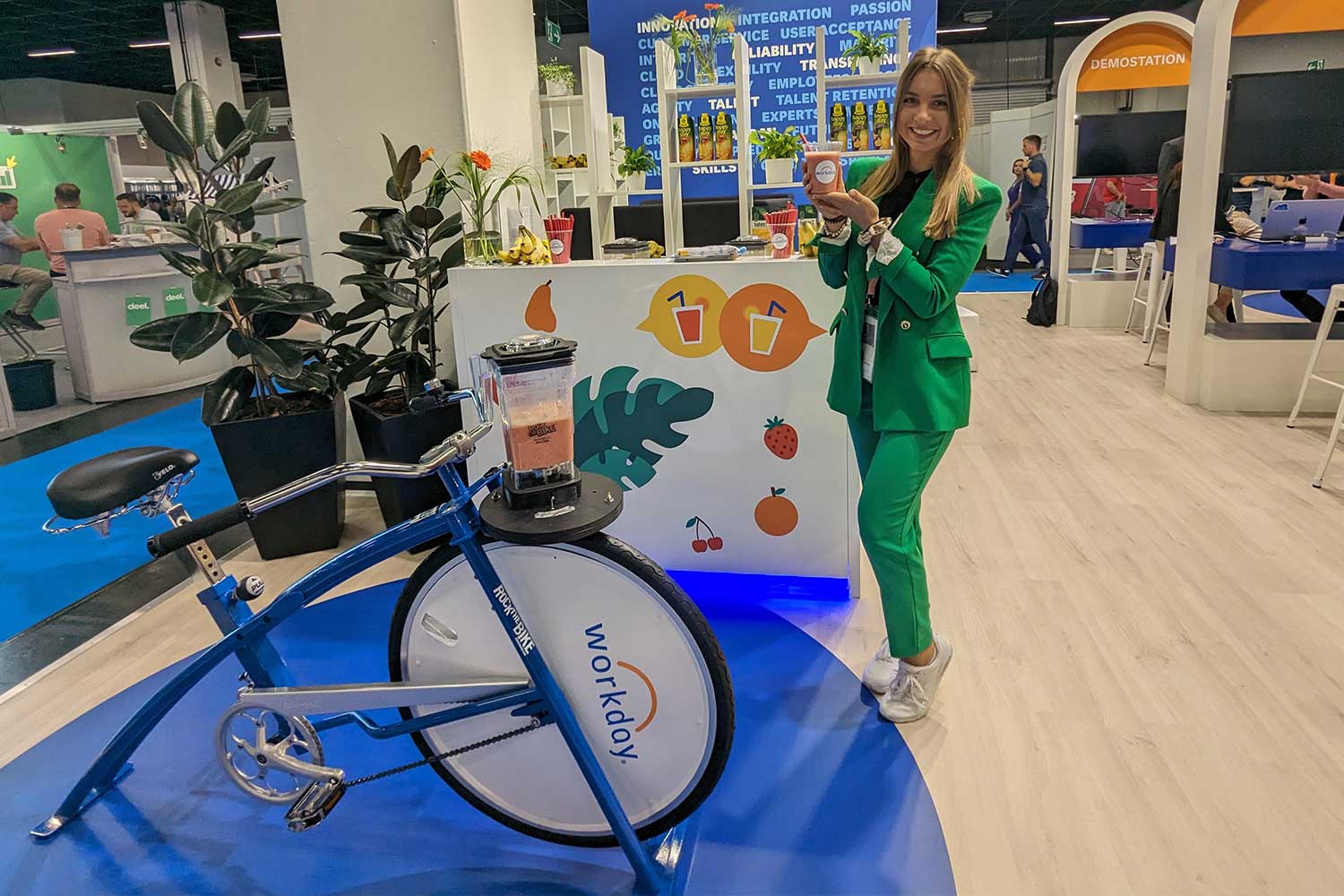 Eine Frau in einem grünen Anzug steht neben einem blauen Smoothie-Bike mit Workday-Branding auf einer Messe in Köln. Sie hält einen frischen Smoothie in der Hand und lächelt in die Kamera. Im Hintergrund sind ein bunter Messestand, Pflanzen und weitere Teilnehmer zu sehen.