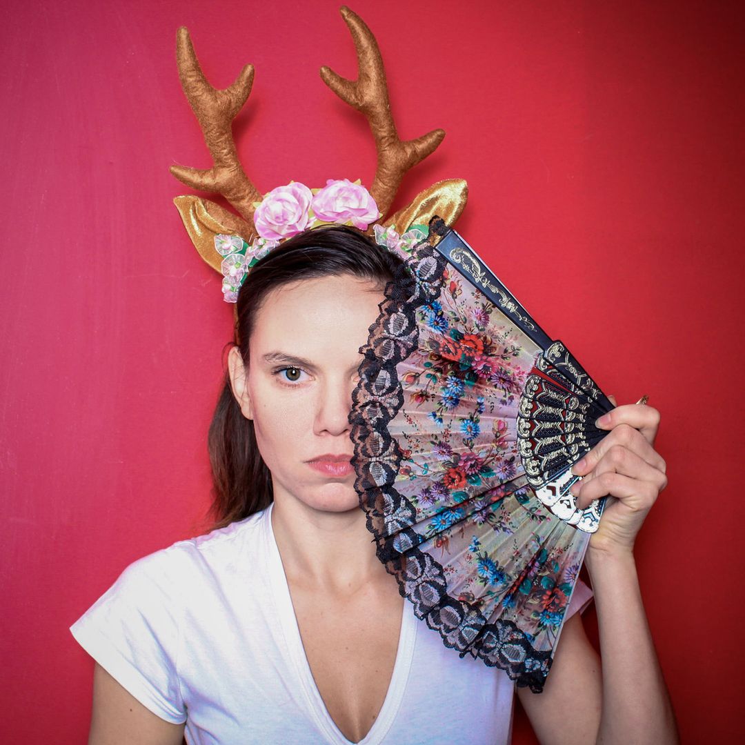Eine Frau trägt ein Geweih-Haarband mit Blumen und hält einen dekorativen Fächer vor ihr Gesicht vor einem roten Hintergrund bei einem Event in Köln.