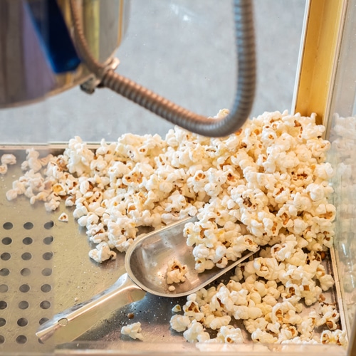 Frisch zubereitetes Popcorn in einer gemieteten Popcornmaschine, sichtbar durch eine gläserne Abdeckung. Eine Metallschaufel liegt auf dem Haufen von warmem, butterigem Popcorn, das gerade aus dem Heizkessel fällt, umgeben von einem silbernen Rahmen und einer klaren Sichtfenster, was den Herstellungsprozess zeigt.