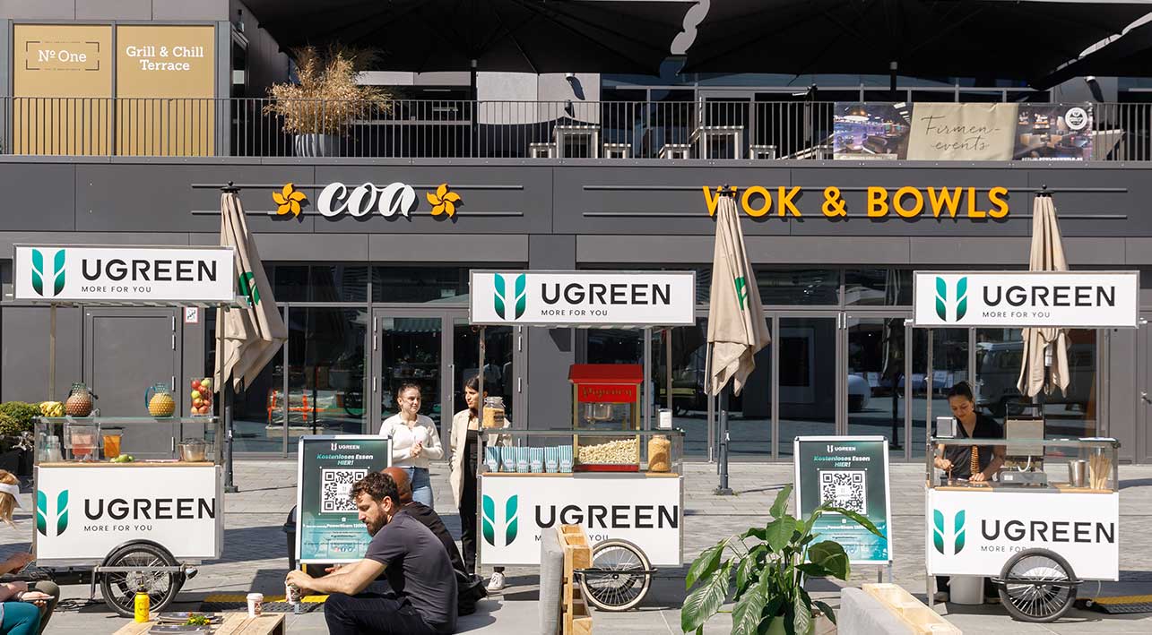 Ein geschäftiges urbanes Setting mit mehreren Promo-Bikes, die für die Marke 'UGREEN' werben. Die Promo-Bikes sind als mobile Verkaufsstände ausgestattet und bieten gesunde Snacks und Getränke an. Menschen unterschiedlicher Altersgruppen genießen die Angebote, während sie in einer sonnigen Fußgängerzone flanieren. Im Hintergrund sind diverse Restaurants und Geschäfte zu sehen.