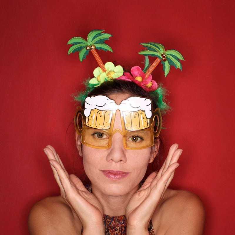 Eine Frau trägt witzige Fotobox-Accessoires wie eine Bierkrug-Brille und ein Palmen-Haarband vor einem roten Hintergrund bei einem Event in Brandenburg.