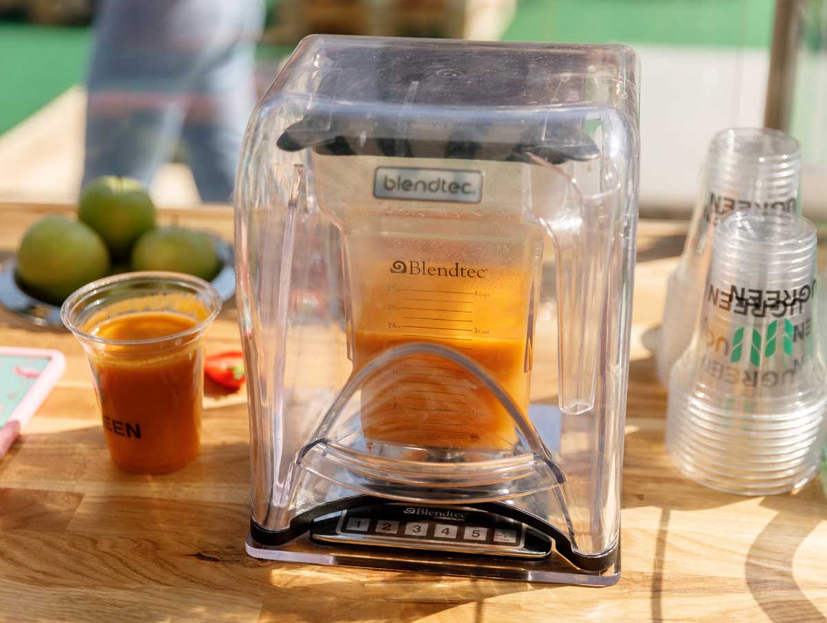 Ein Blendtec-Mixer mixt einen frischen Smoothie neben einem gebrandeten Becher mit dem Logo von UGREEN. Auf dem Tisch liegen grüne Äpfel und weitere Zutaten für Smoothies.