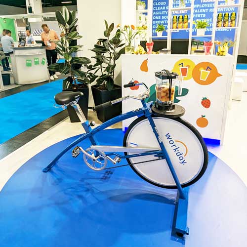 Ein blaues Smoothie-Bike mit dem Branding von Workday steht auf einem Gesundheitstag innerhalb der Firma. Im Hintergrund sind Messestände und Pflanzen zu sehen.