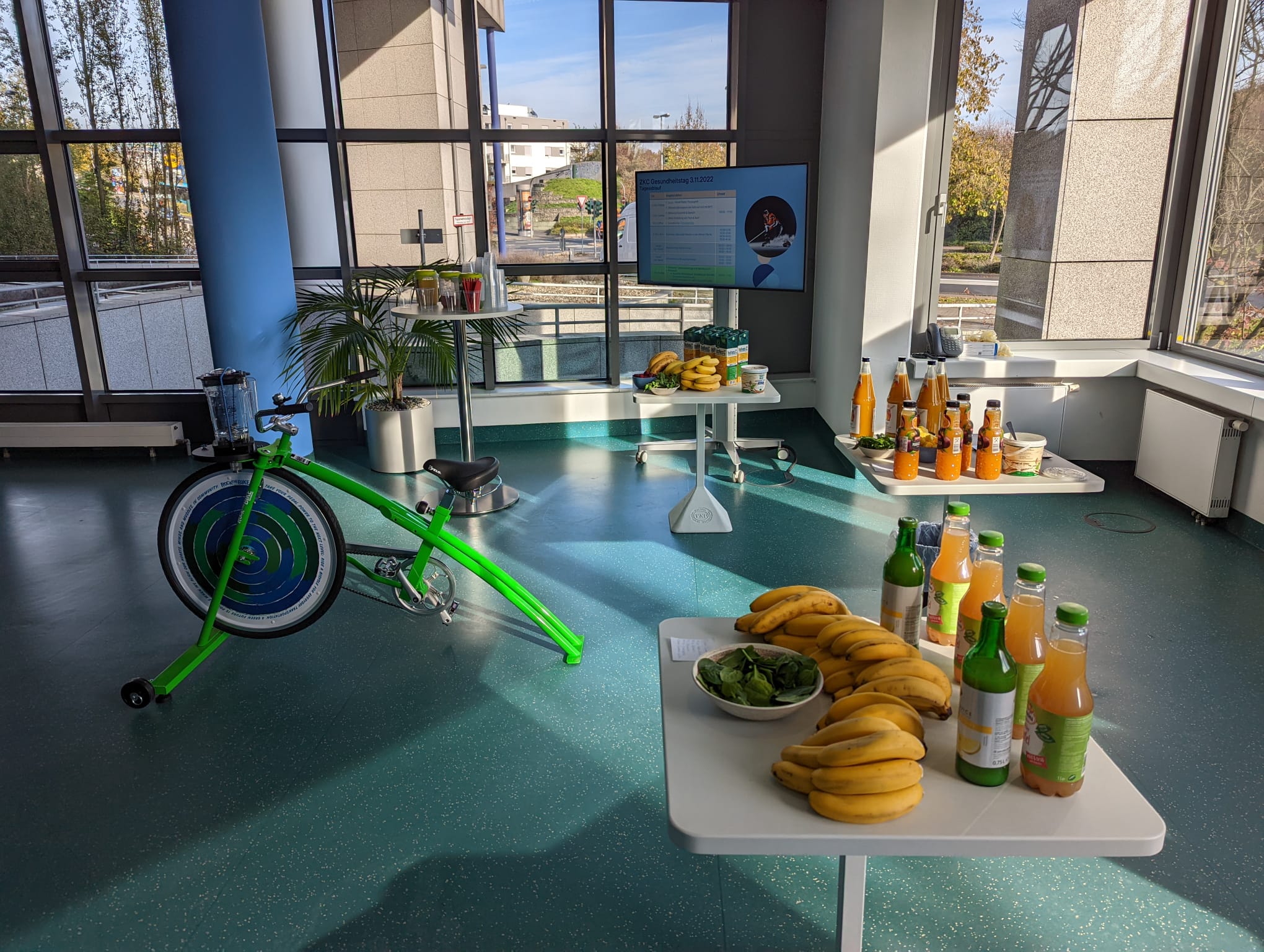 Ein grünes Smoothie-Bike steht in einem modernen Veranstaltungsraum, umgeben von Tischen mit frischen Früchten, Getränken und Smoothie-Zutaten. Im Hintergrund sind große Fenster und ein Bildschirm zu sehen.