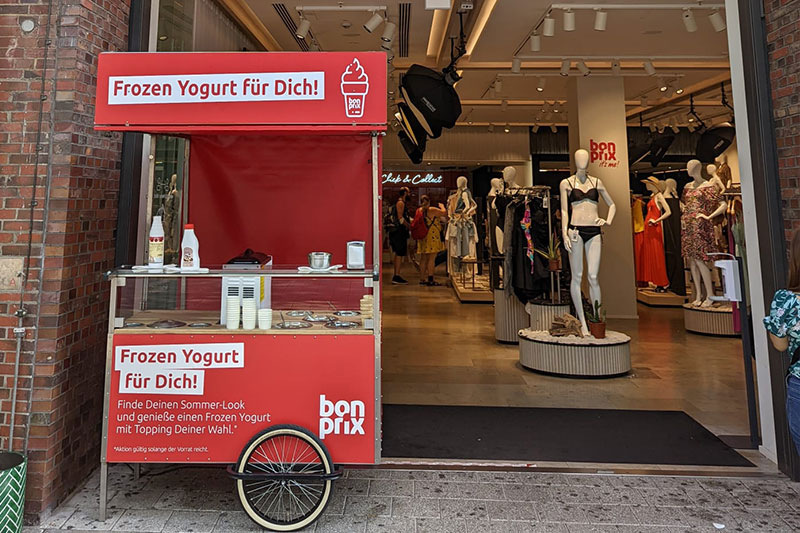Frozen Yogurt Stand vor einem Bekleidungsgeschäft mit Kundenwerbung.