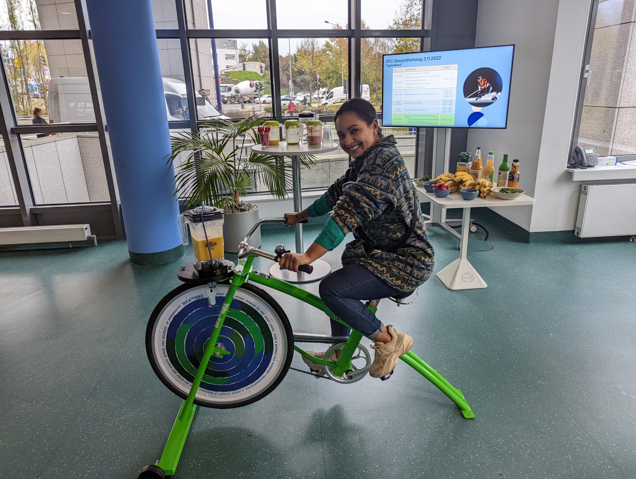 Eine Mitarbeiterin fährt ein grünes Smoothie-Bike und mixt dabei frische Smoothies während eines Gesundheitstags in der Firma. Im Hintergrund sind ein Tisch mit frischen Früchten und Smoothie-Zutaten sowie ein Bildschirm mit dem Tagesablauf des Gesundheitstags zu sehen.