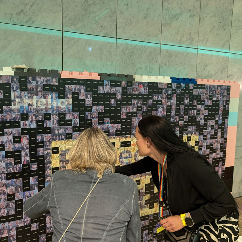 Photo Mosaic Wall Promotion mit Besucherinnen, die ein großes Fotomosaik auf einer Veranstaltung erkunden.